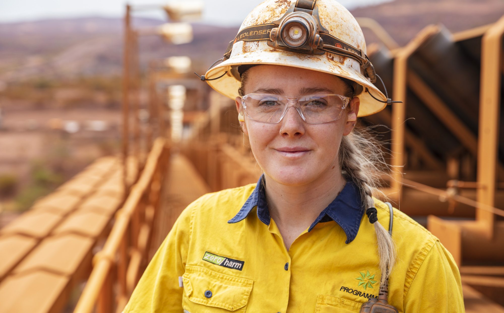 Woman with a soft smile in a yellow safety shirt, safety glasses and hard hat looks into the camera with a mining / operations facility in the background