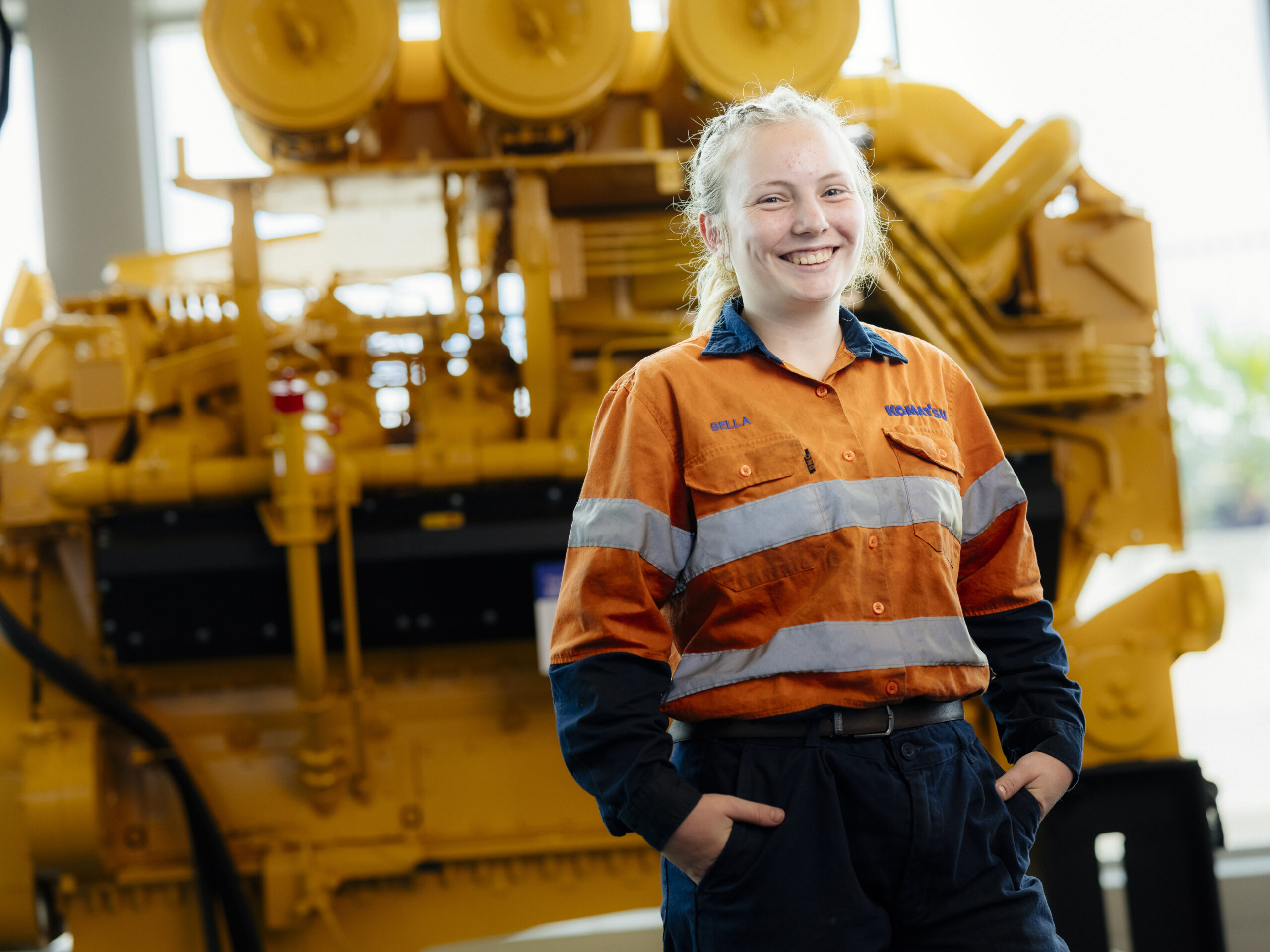 Young woman in orange and blue workwear stands before a large yellow machine